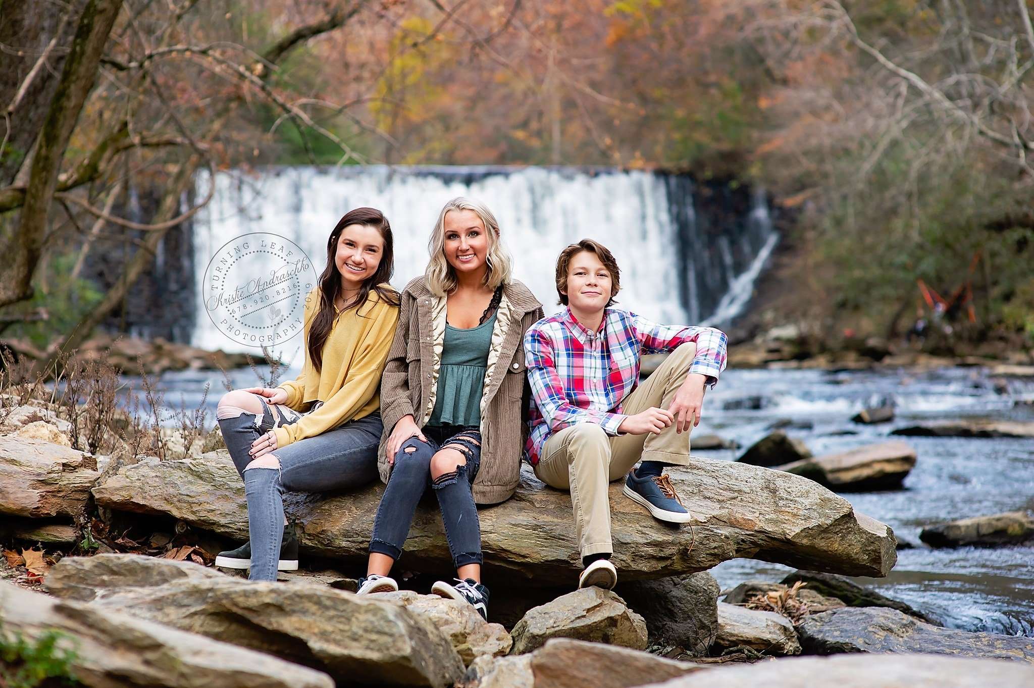 Three people are seated on rocks by a flowing river, with a waterfall and autumn trees in the background. They appear relaxed and happy.Roswell GA at a waterfall