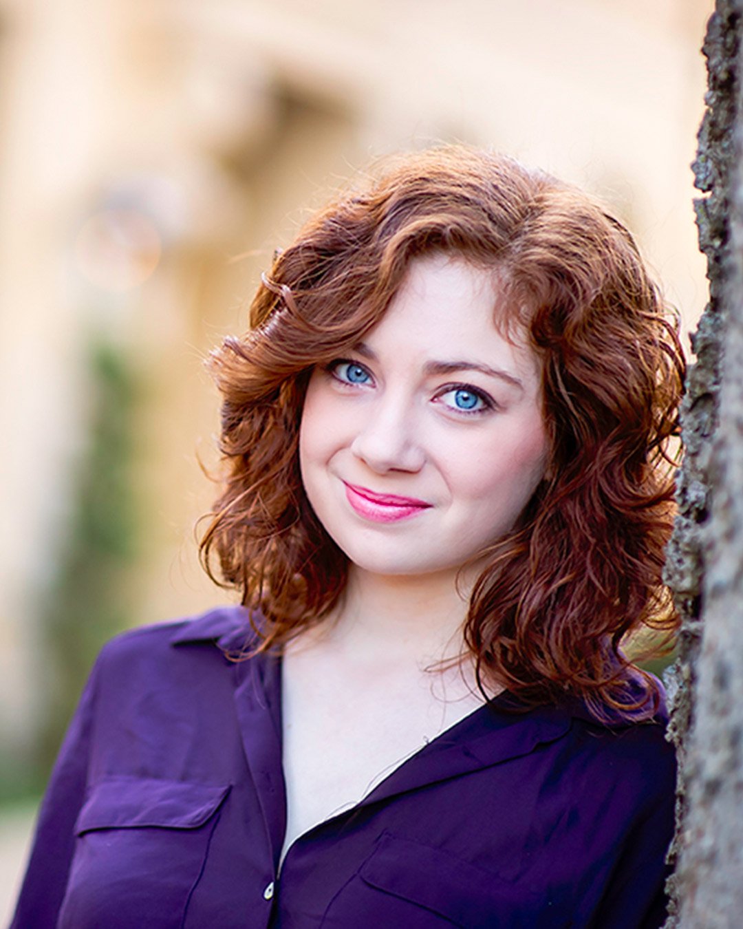 headshots-north-metro-atlanta A woman with curly red hair and blue eyes leans gently against a tree, wearing a purple blouse. She has a slight smile, with a soft, warm background.