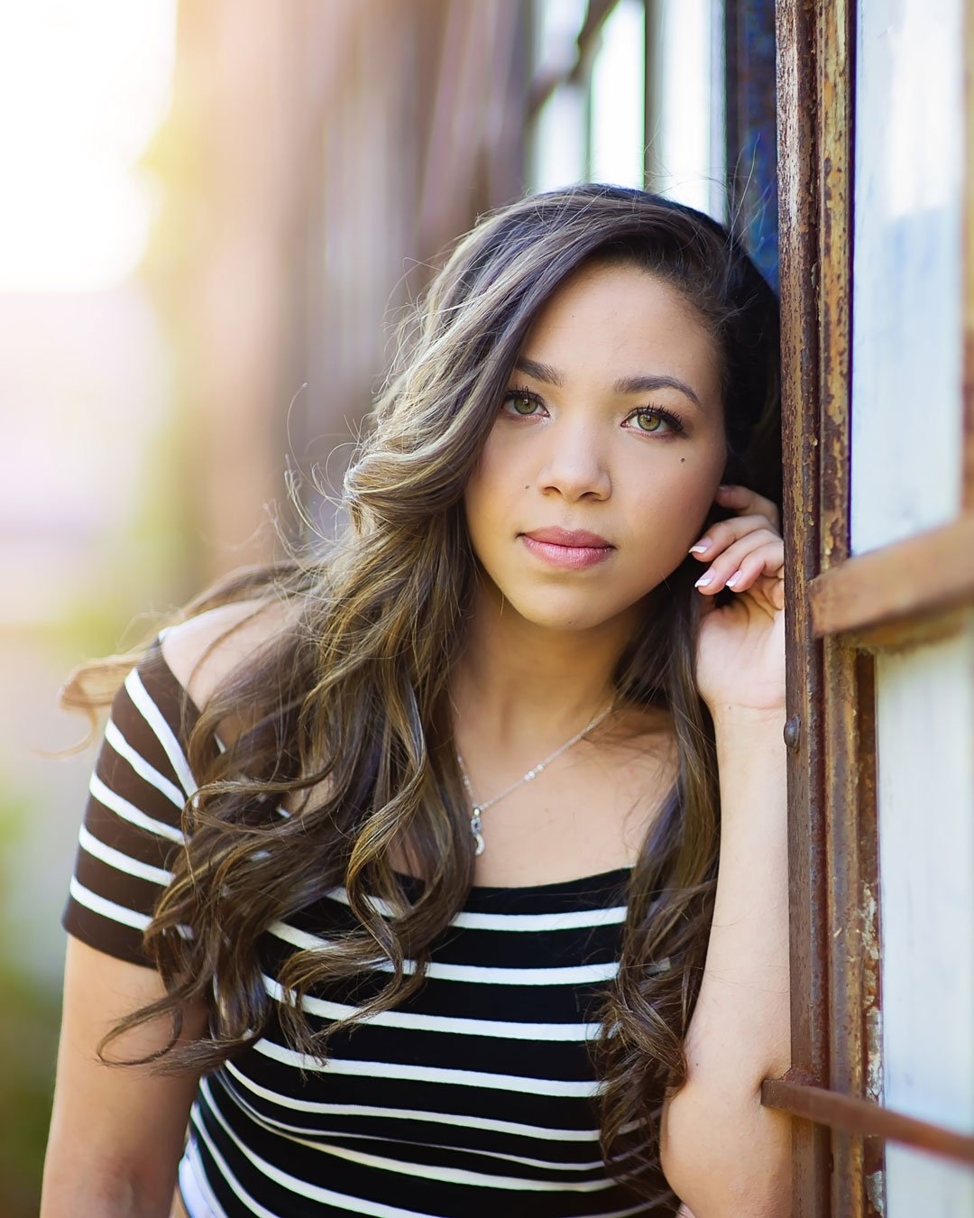 north-metro-atlanta-senior-portrait-session Young woman with long, wavy hair leans thoughtfully against a window, wearing a black and white striped top. Sunlight softly illuminates the scene.