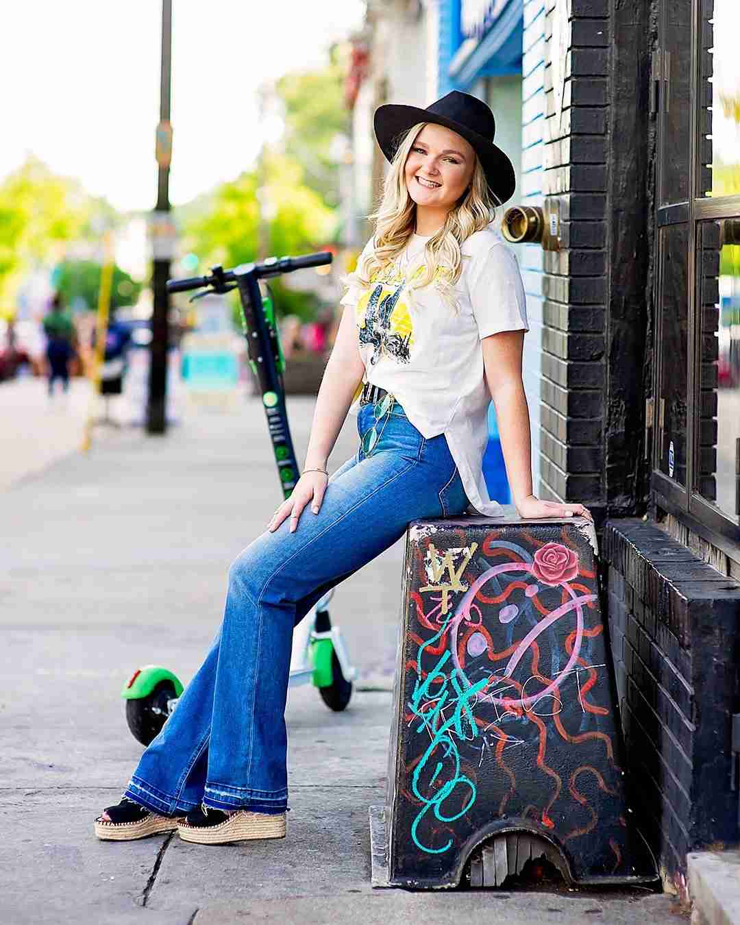 north-metro-atlanta-senior-high-portrait-session Young woman in a black hat and casual outfit leans against a graffitied structure on a city street. A bright green e-scooter is in the background.
