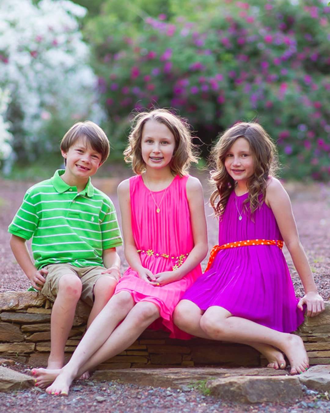 roswell-ga-chilren-portrait-session Three children are sitting on a stone wall in a lush spring garden. Two girls in pink and purple dresses and a boy in a green striped shirt smile warmly.