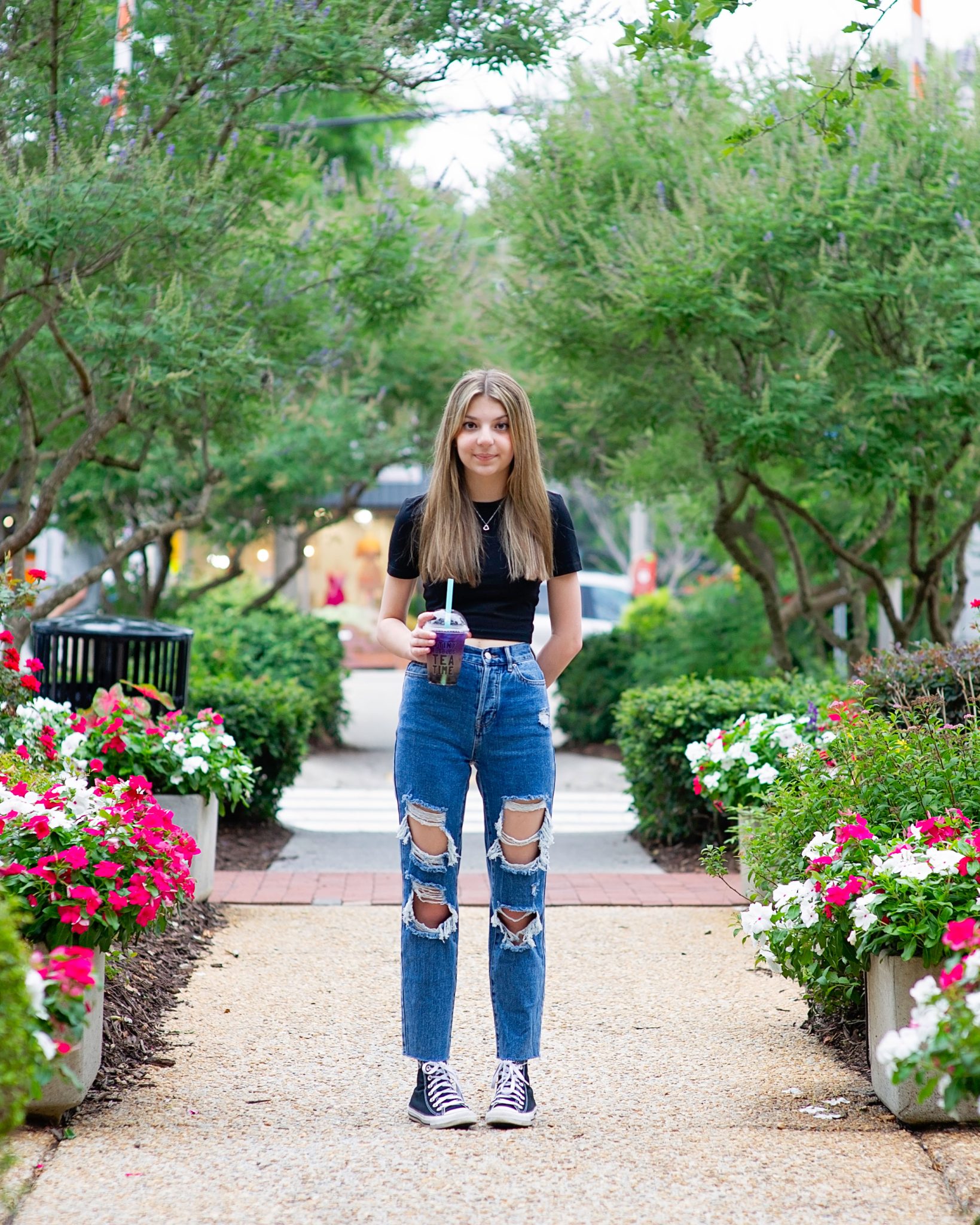 A young woman stands on a garden path lined with flowers, wearing a black top and ripped jeans. She holds a purple drink, smiling softly, conveying a relaxed vibe.