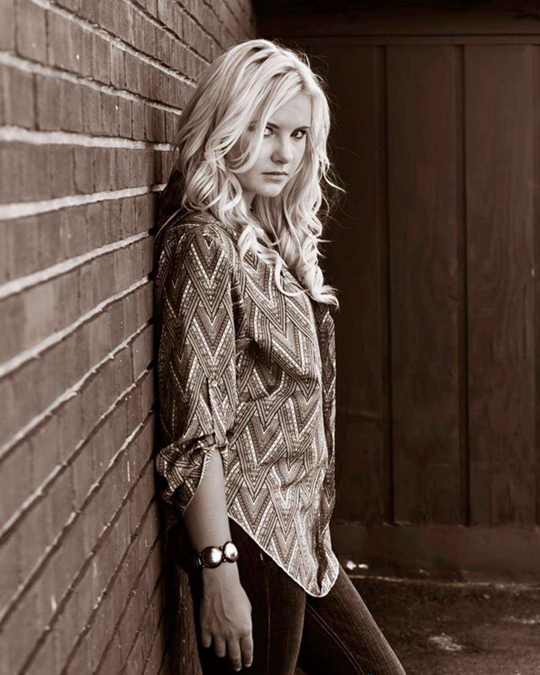 woodstock-ga-creative-editorial-senior-pictures A woman with long blond hair leans against a brick wall, wearing a patterned blouse and bracelet. The tone is contemplative and neutral.