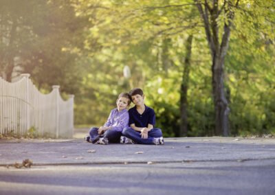 Two young boys sit on a road, leaning against each other, surrounded by autumn trees. The scene is serene with soft, golden light.