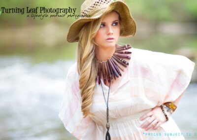 A woman in a flowy white dress and wide-brimmed straw hat poses by a blurred river. She wears a large wooden necklace, exuding a bohemian vibe.
