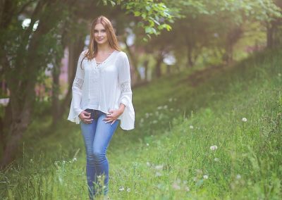A woman in a white blouse and jeans stands smiling in a sunlit, lush green meadow surrounded by trees. The scene conveys a serene, cheerful mood.