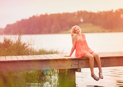 A girl in a pink dress sits joyfully on a wooden dock by a tranquil lake, with soft sunlight and green trees in the background, conveying a serene, summer vibe.