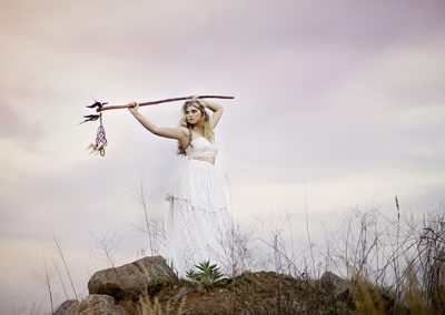 A woman in a flowing white dress stands atop rocks, holding a decorated staff against a pastel pink and blue sky, evoking a mystical, serene vibe.