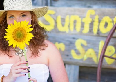 Woman in a straw hat holds a sunflower, smiling slightly. Background shows rustic wood with a painted "Sunflowers for Sale" sign. Bright and cheerful mood.