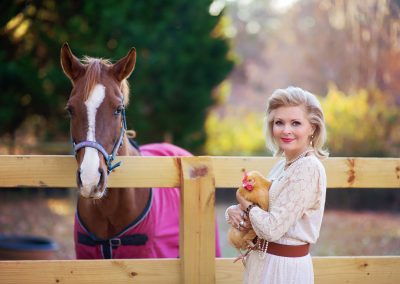 A woman in a cream dress holds a chicken, standing near a brown horse with a pink blanket by a wooden fence. The scene is warm and serene.