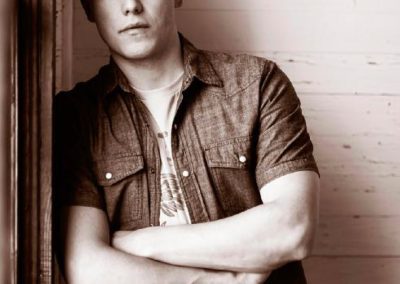 Sepia-toned photo of a person leaning against a wall, arms crossed, wearing a denim shirt. The expression is serious and contemplative.