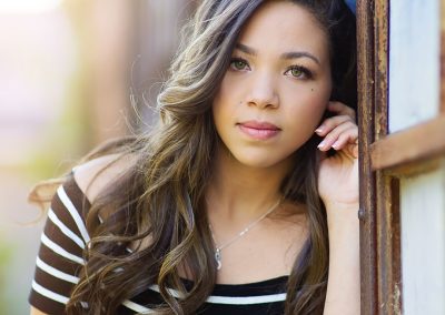 Young woman with long, wavy hair leans thoughtfully against a window, wearing a black and white striped top. Sunlight softly illuminates the scene.