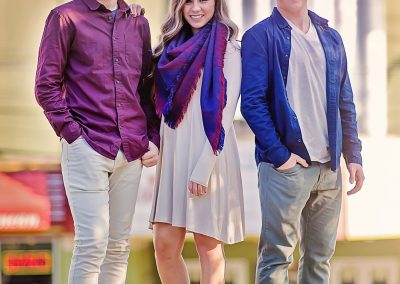 Three people stand outdoors, smiling. The woman in the center wears a white dress with a blue scarf, flanked by men in casual purple and blue shirts. Bright, sunny ambiance.