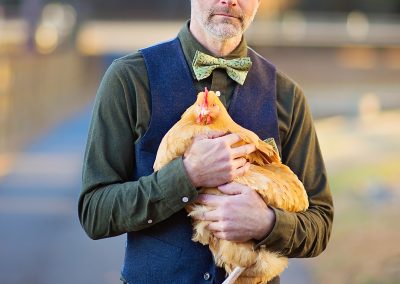 A man stands holding a chicken, wearing a green shirt, blue vest, and bow tie. The background is softly blurred with autumn colors, conveying a calm mood.