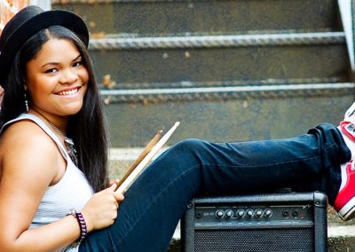 A smiling person sits on outdoor steps, holding drumsticks, leaning against a small amp. They wear a black hat and red sneakers, exuding a relaxed vibe.