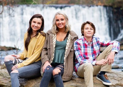 Three people sit on rocks in front of a waterfall. They are smiling, dressed casually in fall attire, conveying a warm and relaxed atmosphere.