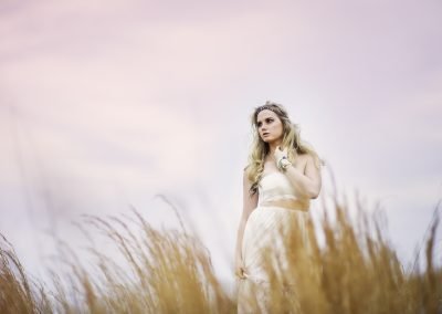 A woman in a flowing white dress stands in a field of tall grasses at sunset. The sky glows in soft pink hues, creating a serene, dreamy atmosphere.