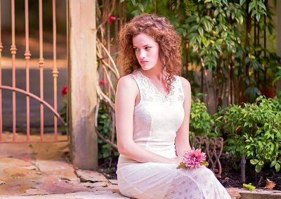 A woman in a white lace dress sits on stone steps, holding pink flowers, near a wrought iron gate. A straw hat rests beside her, conveying a serene mood.