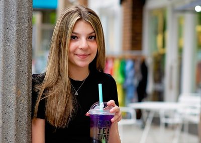 A young woman with long brown hair smiles while holding a purple bubble tea. She leans against a pole, wearing a black top and jeans. Outdoor cafe setting.