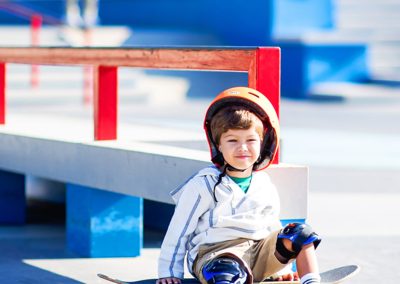 A young boy sitting on a skateboard at a skate park, wearing a helmet and knee pads. He's smiling, with a colorful ramp and rail in the background.