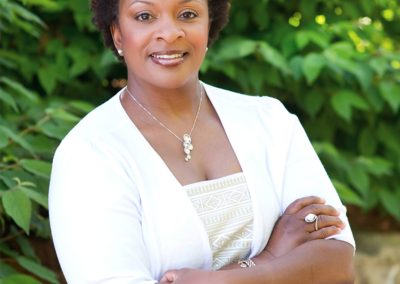 Smiling woman with short curly hair, wearing a white outfit and silver jewelry, stands confidently with arms crossed against a lush green leafy background.