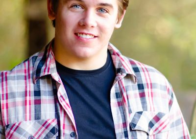 Young man with slicked-back hair, wearing a plaid shirt over a black tee, smiles warmly at the camera. Sunlight creates a soft, natural background.