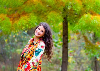 Young woman with long curly hair in a colorful coat smiles brightly against a backdrop of vibrant autumn trees, conveying joy and warmth.
